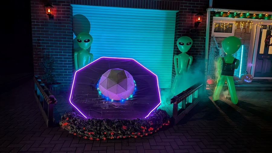 Wide view of the crashed UFO. It is octagonal with an illuminated geodesic dome in the centre. The edge is also illuminated with an LED strip. To simulate an impact site, at the base of the UFO there is expanding foam with red LEDs embedded within. Two inflatable aliens stand in the background.